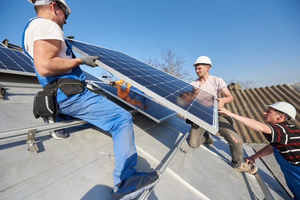 Male team engineers installing stand-alone solar photovoltaic panel system. Electricians lifting blue solar module on roof of modern house. Alternative energy ecological concept.